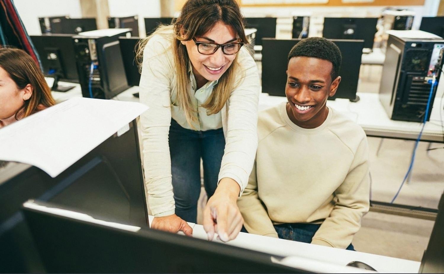 Teacher pointing at a computer monitor helping a student in a school computer lab